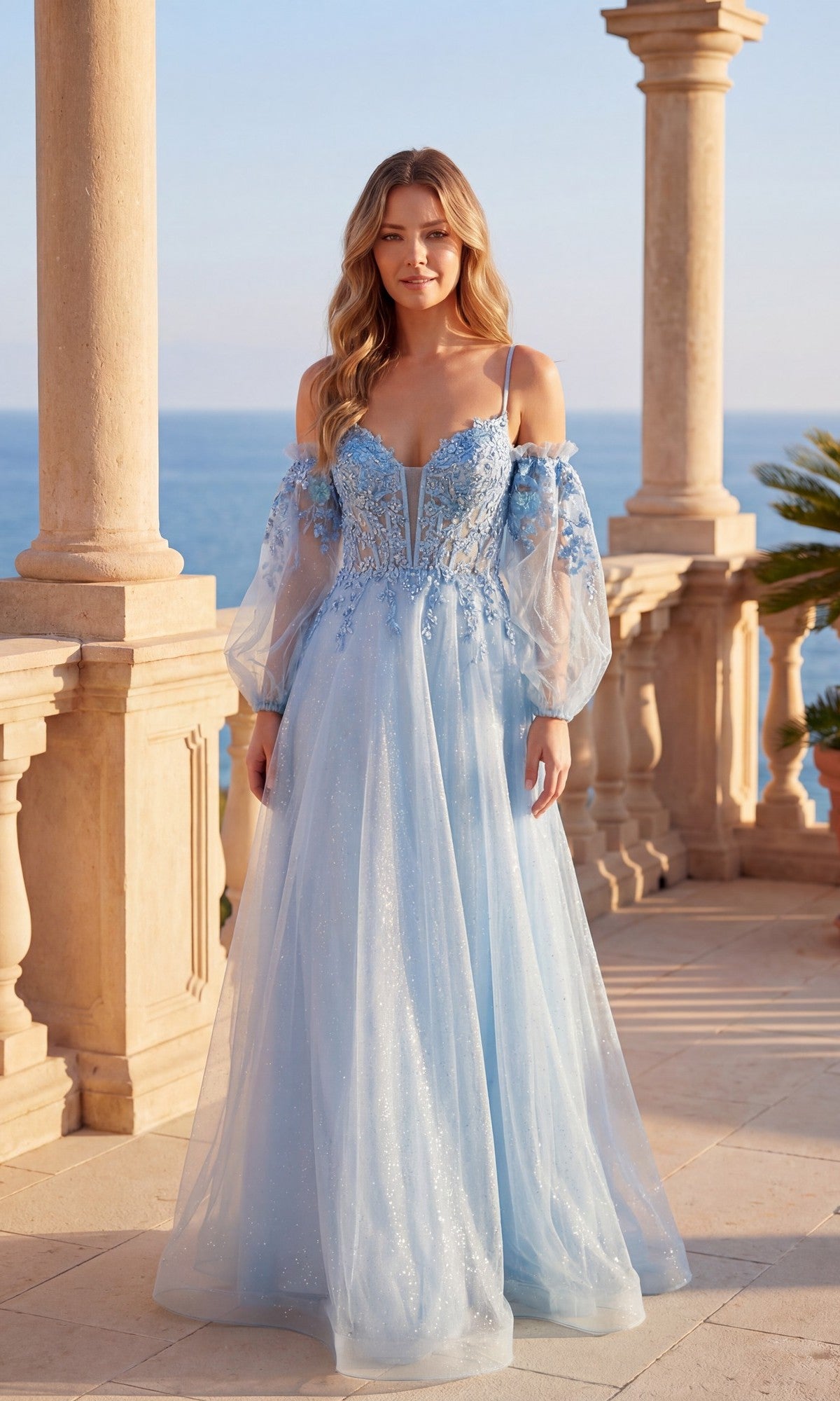 A woman in the Chicas C358 long formal dress in sky blue, featuring an off-shoulder corset bodice and floral embroidery, stands on a stone terrace overlooking the ocean.